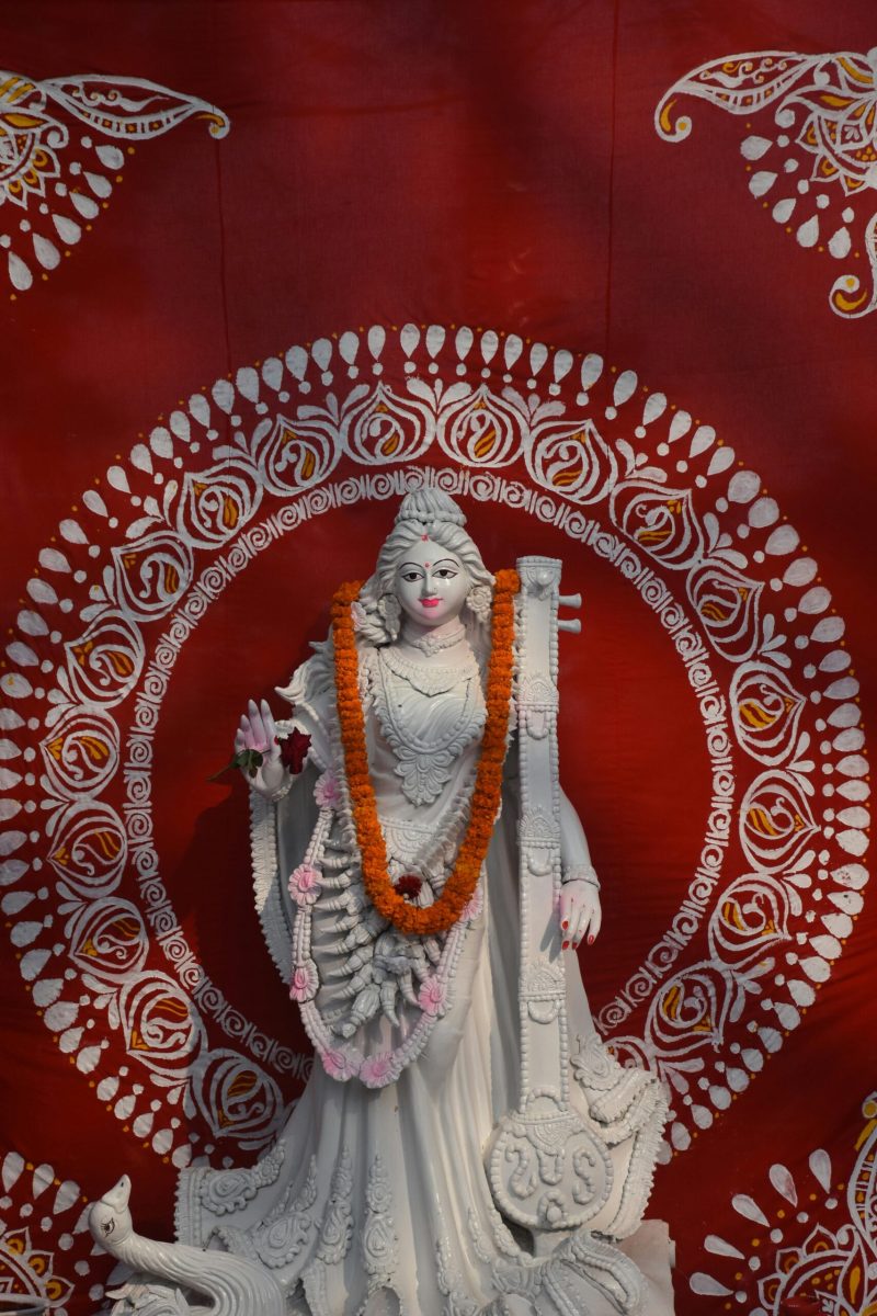 Elegant statue of Hindu goddess Saraswati garlanded against a vibrant red backdrop.